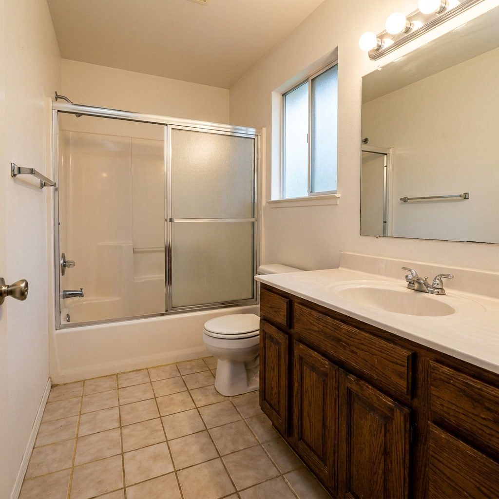 Before: Dated 1990s bathroom with chrome sliding glass tub-shower doors, beige ceramic tile floor, oak vanity, and old Hollywood light bar in Delray Beach, FL
